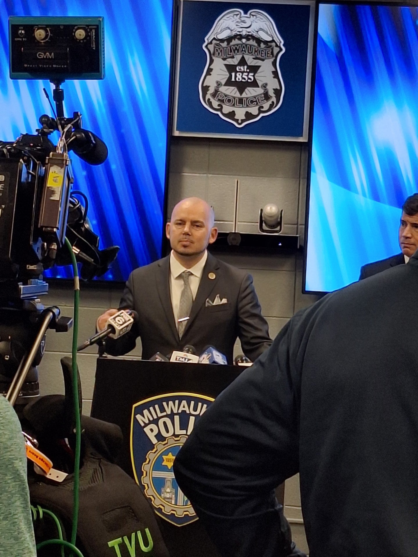 A man in a suit speaks at a podium with Milwaukee Police logos, surrounded by cameras and microphones at a press conference.