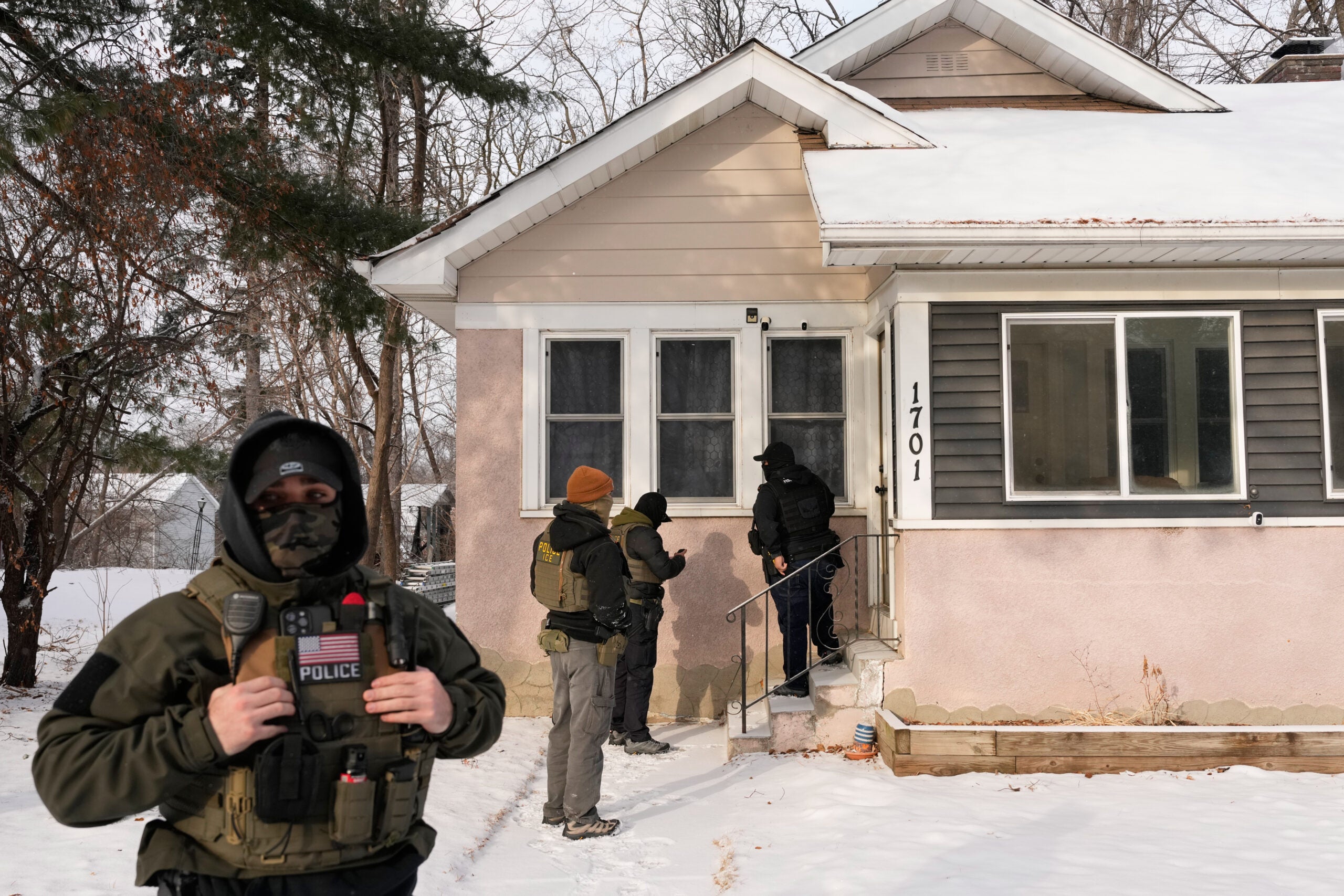 Four armed law enforcement officers conduct a raid at the front door of a house in winter; one officer stands in the foreground wearing a vest labeled POLICE.