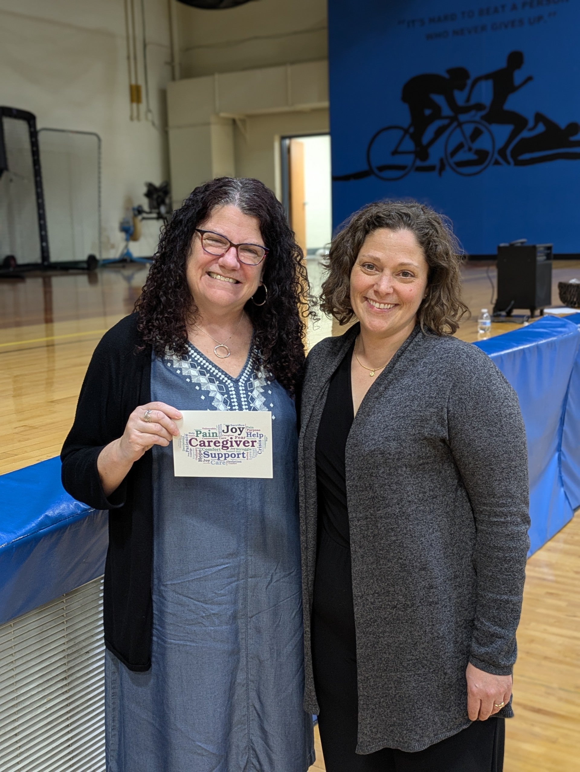 Two women stand side by side in a gymnasium; one holds a sign reading Joy Caregiver Support. A blue wall with a cyclist mural is visible in the background.