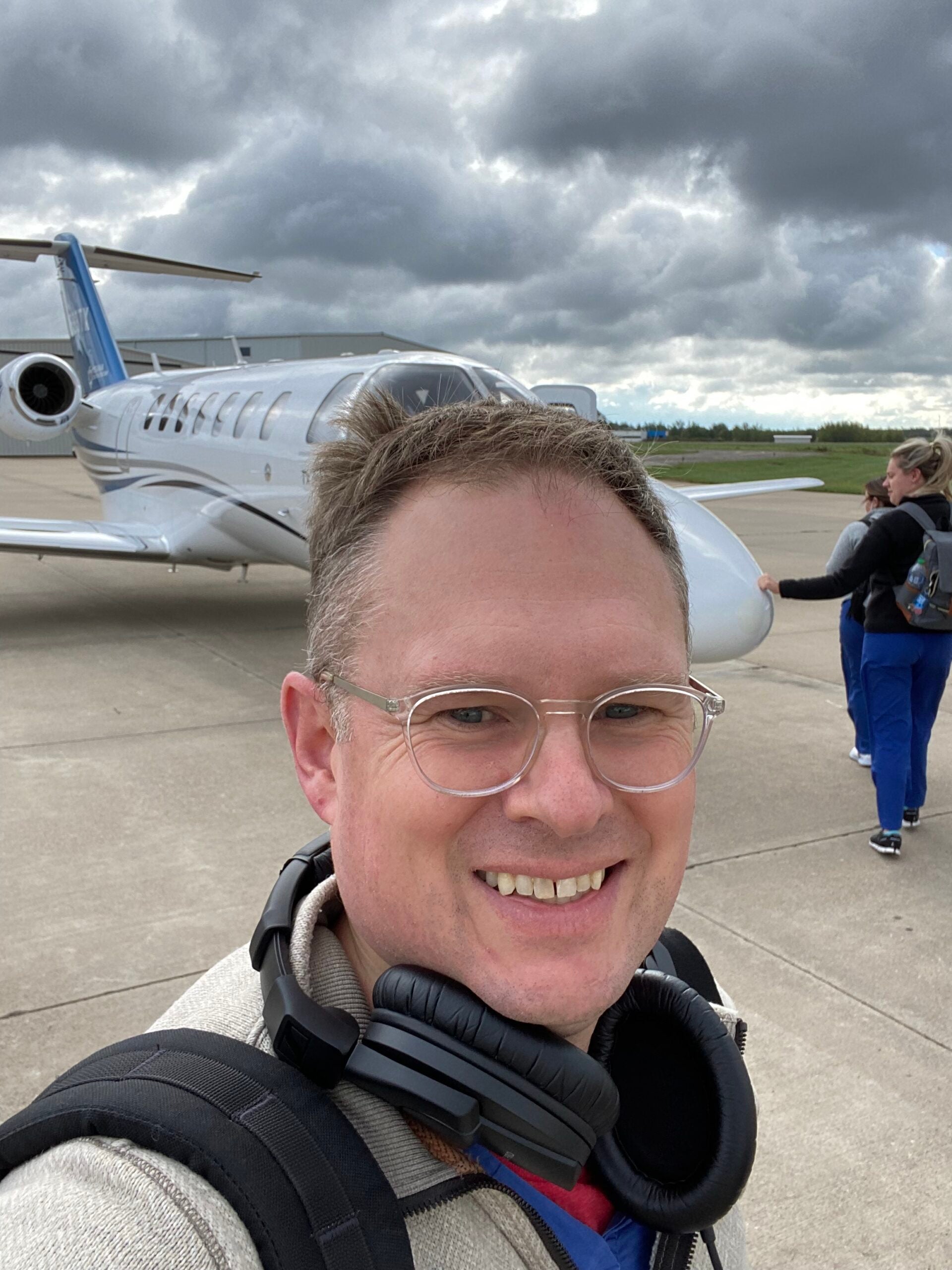 A man with glasses and headphones around his neck smiles for a selfie on an airport tarmac in front of a small private jet.