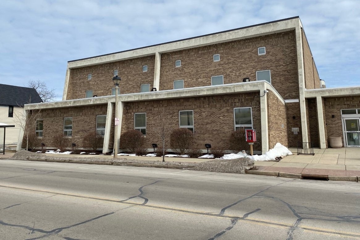 A large, rectangular brick building with multiple small windows, a sidewalk in front, patches of snow, and a red public phone booth near the entrance.