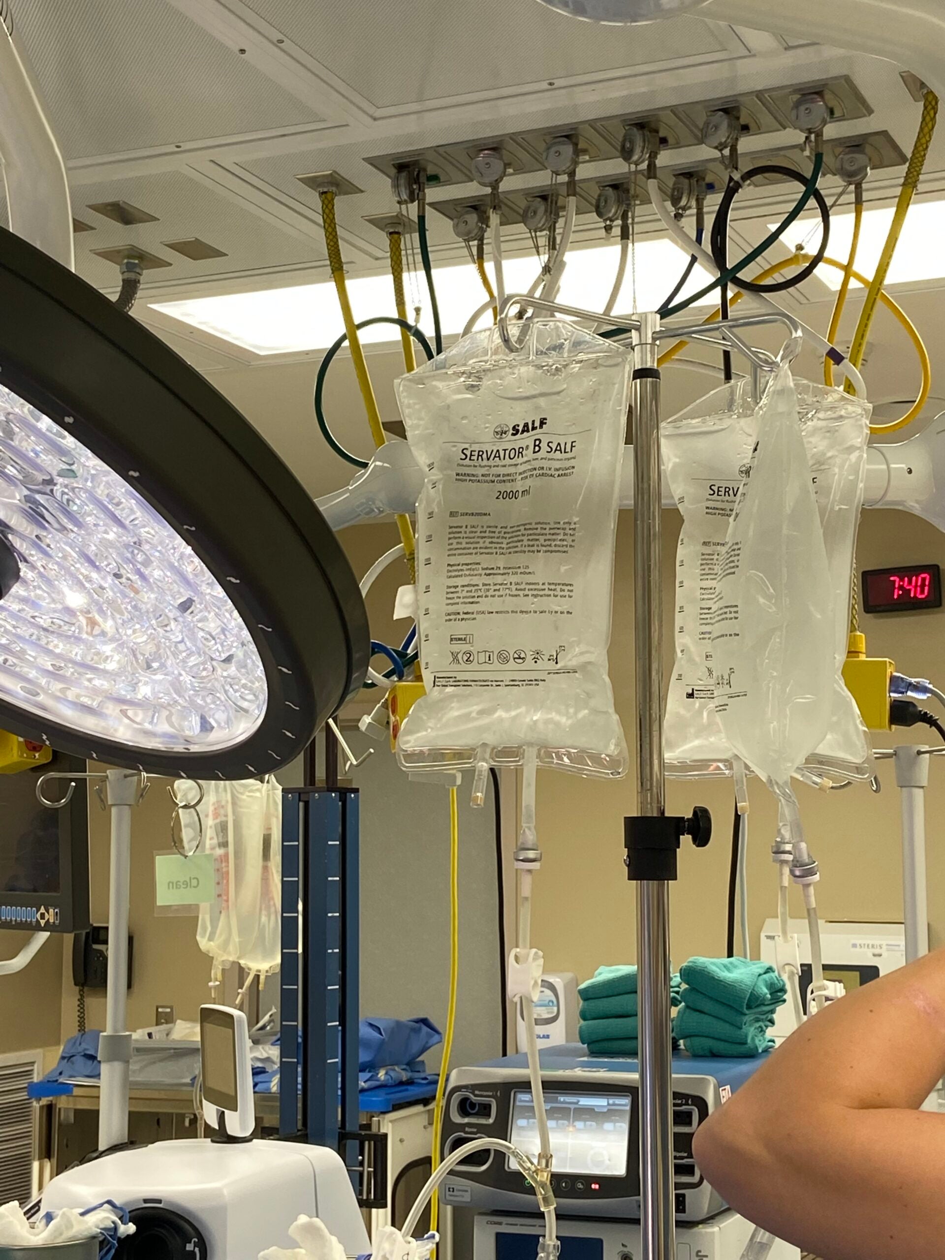 IV saline bags and medical equipment are set up in a hospital operating room, with a surgical light and monitors visible in the background.